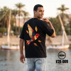 Man looking back near the Nile riverbank wearing a black DEDO oversized t-shirt with a colorful geometric bird back graphic, model V2B-009.